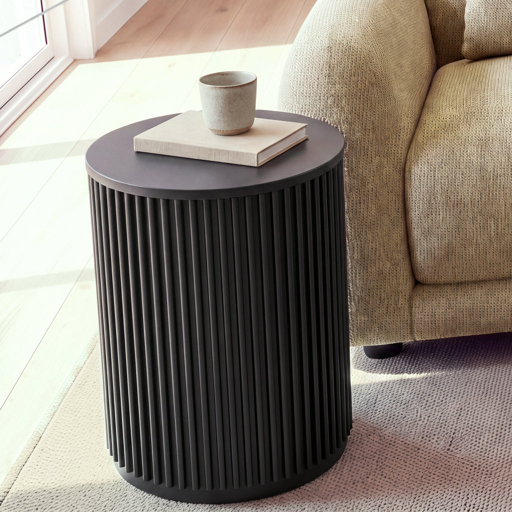Ayla Fluted 40cm Round Side Table Black beside the arm of a light sofa, with a cup on a linen book, in a sunlit living room