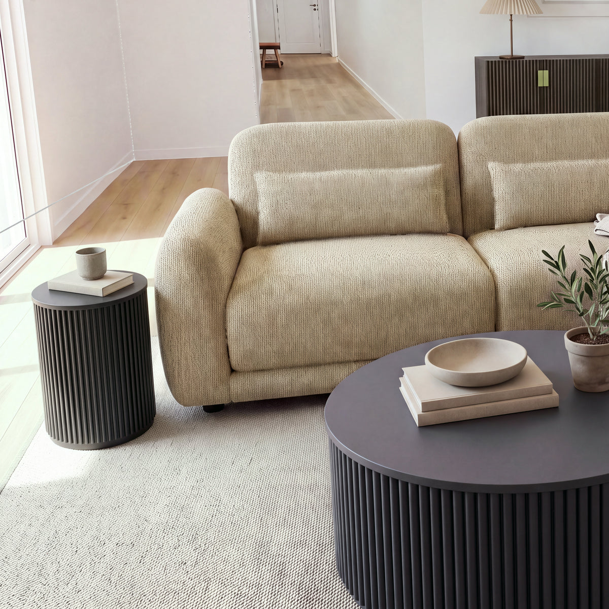 Ayla Fluted 40cm Round Side Table Black beside the arm of a modular sofa, with a round black fluted coffee table in front and a hallway behind