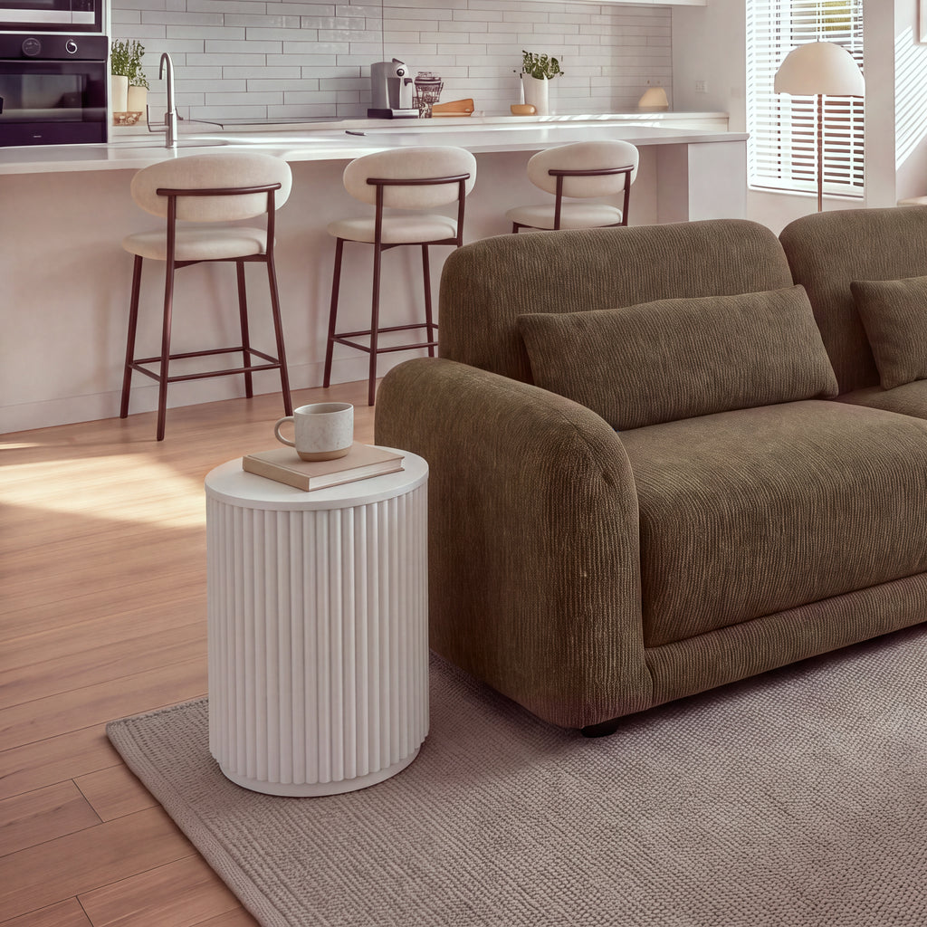 Ayla Fluted 40cm Round Side Table White beside a sofa in an open-plan living room with a kitchen and bar stools behind, a mug on a book on top