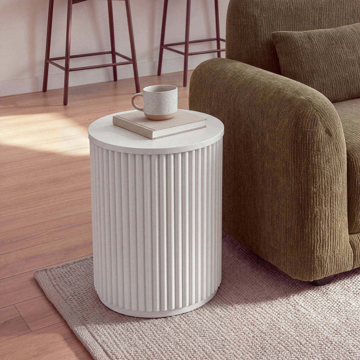 Ayla Fluted 40cm Round Side Table White beside the arm of a sofa on a rug, closer view with a mug on a book