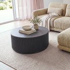 Ayla Fluted 90cm Round Coffee Table Black beside a sofa chaise near glass doors, with a bowl on books and a potted olive tree