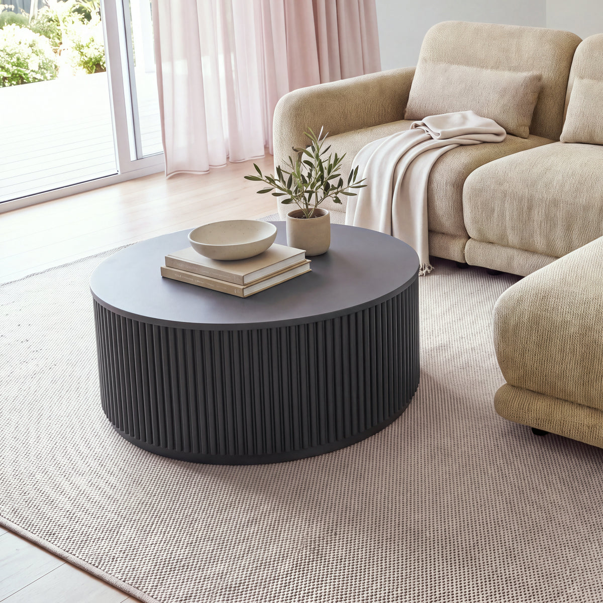 Ayla Fluted 90cm Round Coffee Table Black beside a sofa chaise near glass doors, with a bowl on books and a potted olive tree