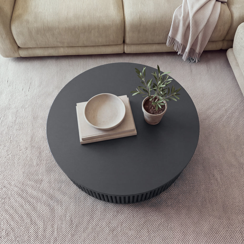 Ayla Fluted 90cm Round Coffee Table Black seen from above on a rug in front of a sofa, with a bowl on books and a small potted olive tree