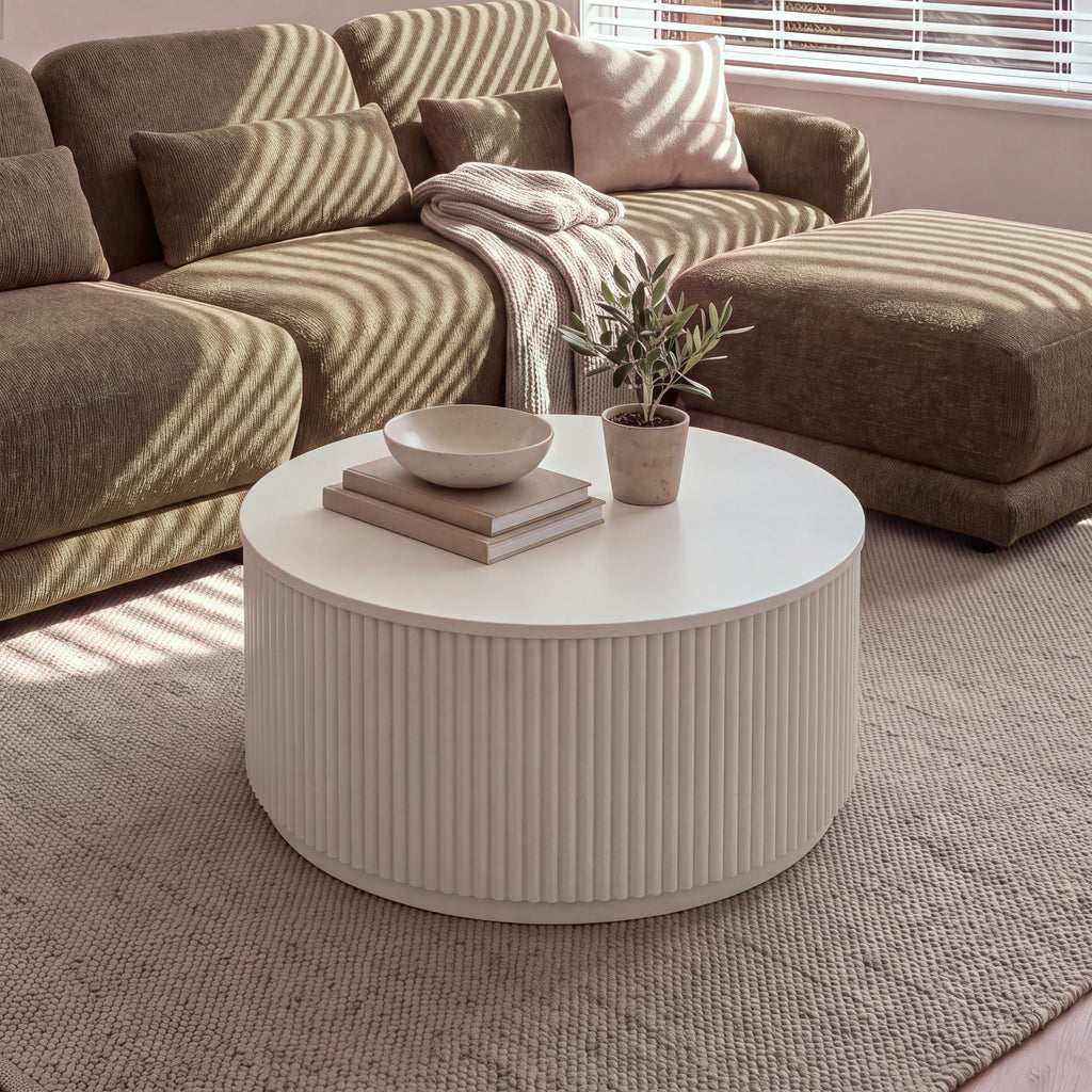 Ayla Fluted 90cm Round Coffee Table White in front of a modular sofa in striped sunlight, with a bowl, books and a potted olive tree on top