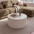 Ayla Fluted 90cm Round Coffee Table White in front of a modular sofa in striped sunlight, with a bowl, books and a potted olive tree on top