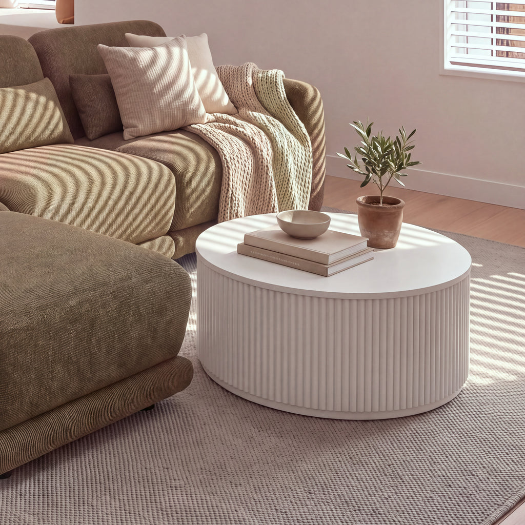 Ayla Fluted 90cm Round Coffee Table White beside a sofa chaise, with a bowl on stacked books and a potted olive tree