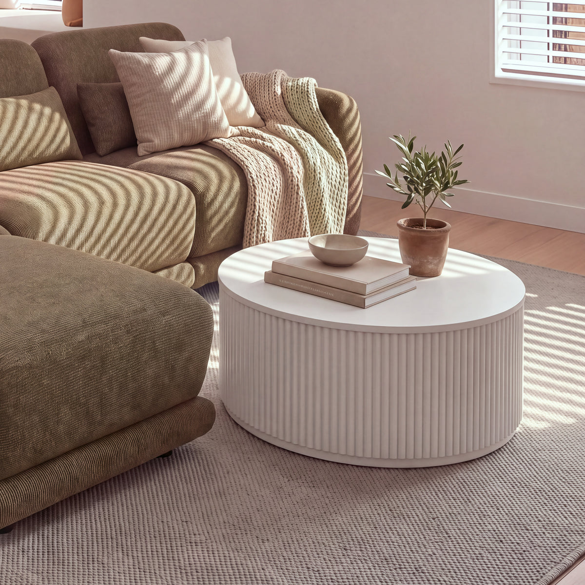 Ayla Fluted 90cm Round Coffee Table White beside a sofa chaise, with a bowl on stacked books and a potted olive tree