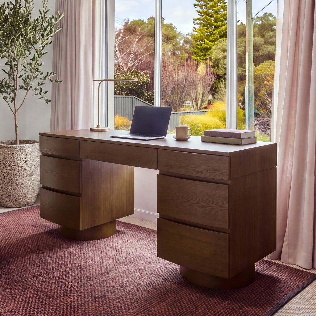 Dorit 172cm 7 Drawer Home Office Desk Walnut in a home office in front of a garden window, with a laptop and a desk lamp on top