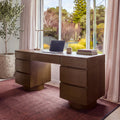 Dorit 172cm 7 Drawer Home Office Desk Walnut in a home office in front of a garden window, with a laptop and a desk lamp on top