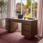 Dorit 172cm 7 Drawer Home Office Desk Walnut in a home office in front of a garden window, with a laptop and a desk lamp on top