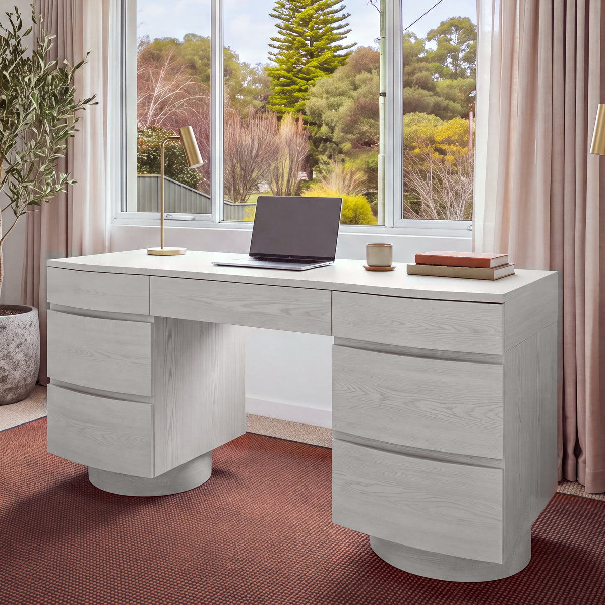 Dorit 172cm 7 Drawer Home Office Desk White in a home office in front of a garden window, with a laptop and a desk lamp on top