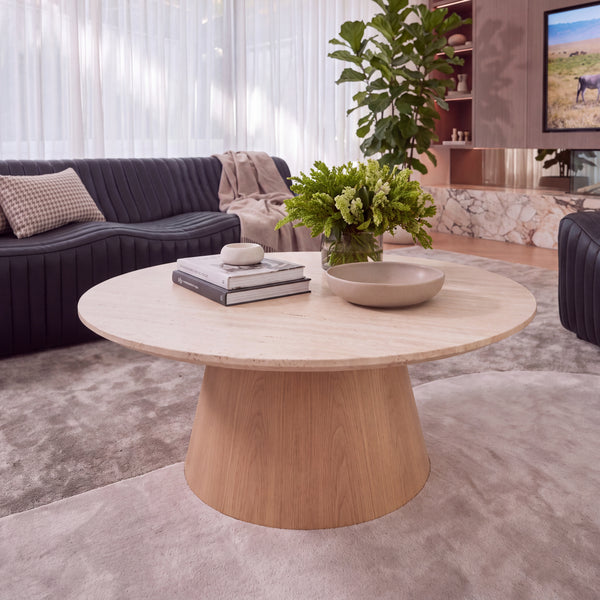 Hera 100cm Round Coffee Table Natural, in a living room beside a dark sofa, styled with flowers and books