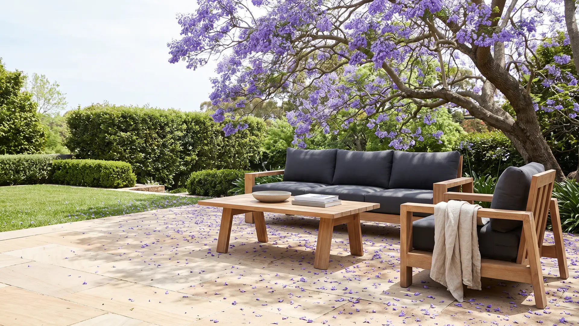 Stud outdoor sofa, armchair and coffee table in natural timber on a sunny terrace under a flowering jacaranda